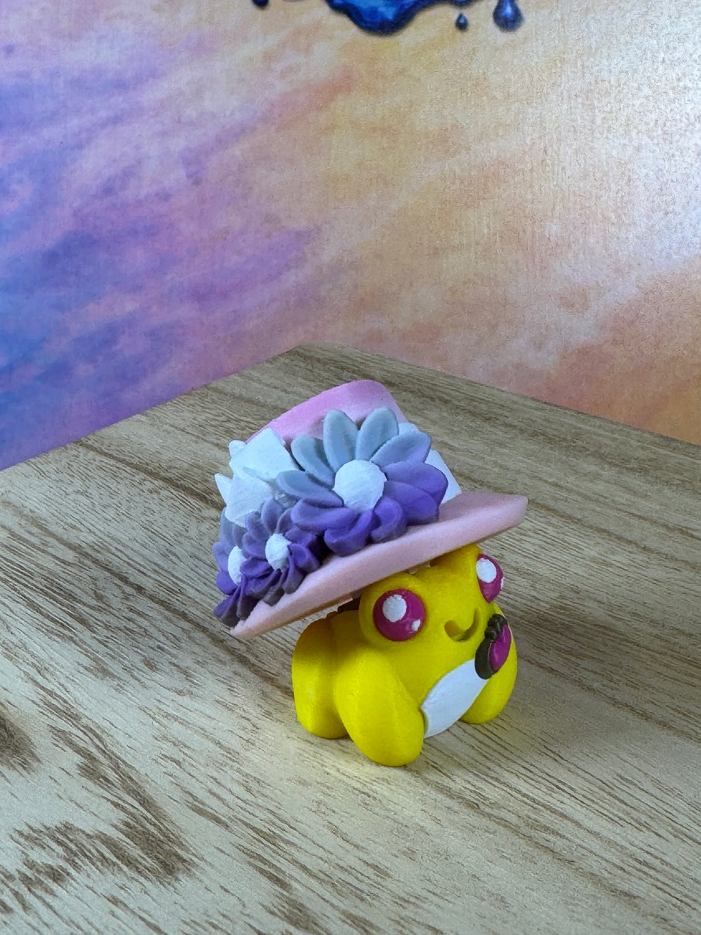 Lady Gilded Frog Clicker, yellow frog wearing a pink floral hat with purple flowers